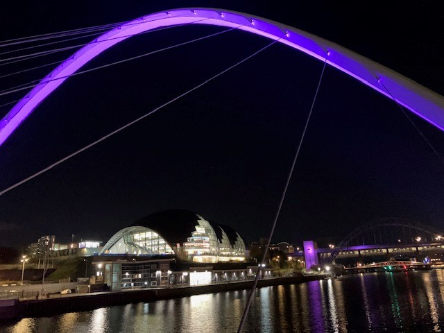 The image shows the Millennium Bridge across the River Tyne at night, lit up with purple light. The Glasshouse can be seen in the background