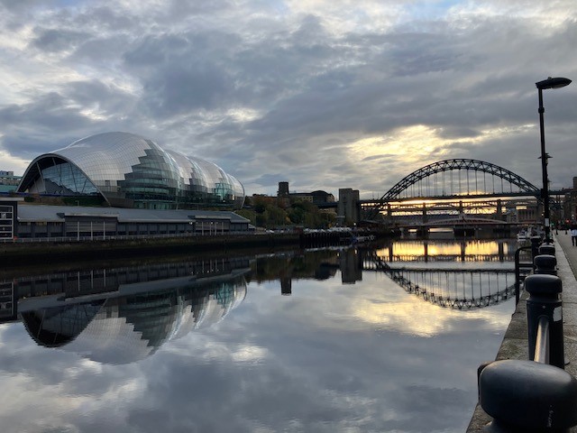 The image shows the River Tyne in the foreground with The Glasshouse and Tyne Bridge in the background, and reflected in the water
