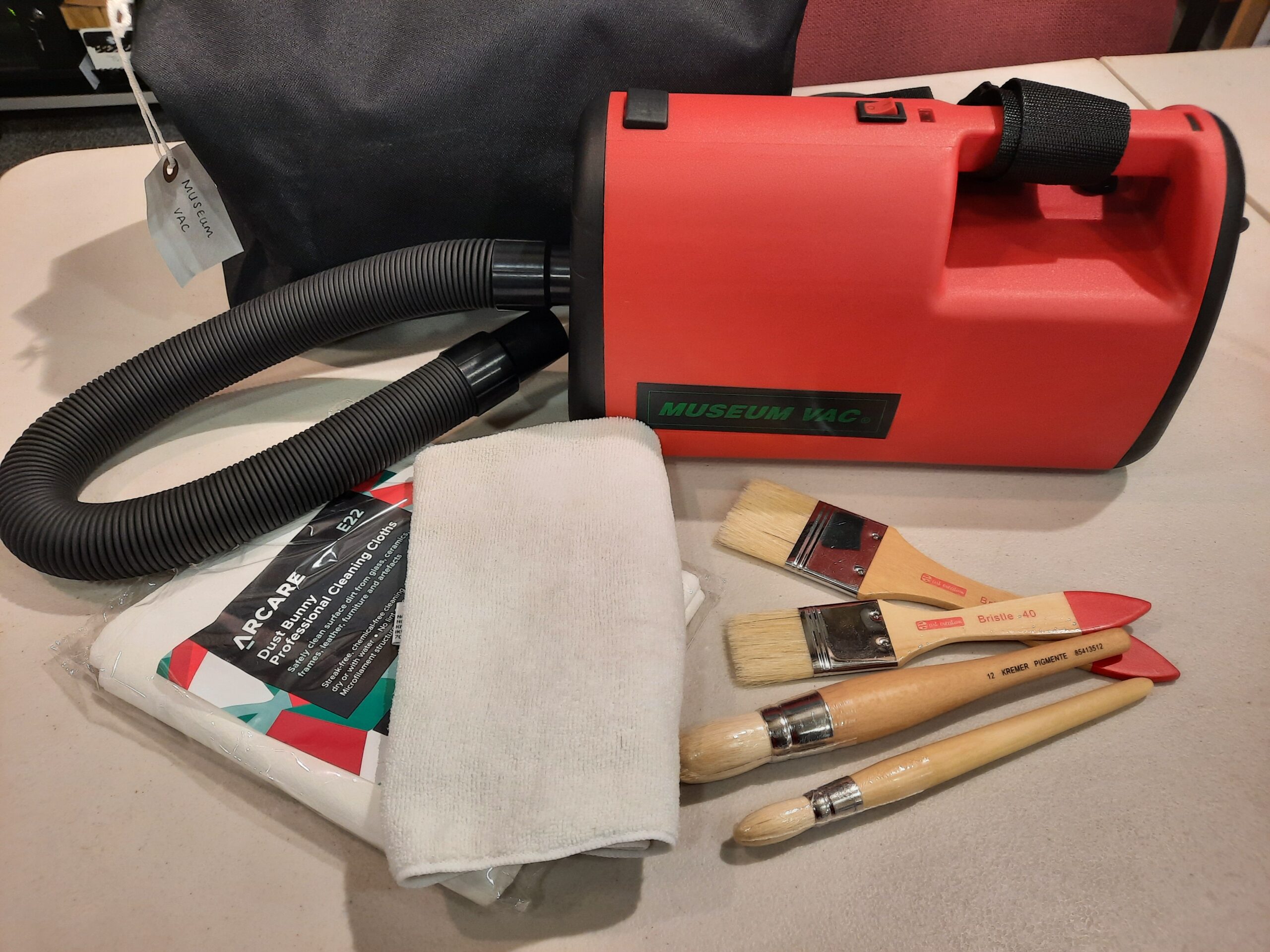 The image shows museum cleaning equipment including a red museum vacuum cleaner, assorted cleaning brushes, a white microfibre cloth and some Dust Bunny Professional cleaning cloths used for different types of museum items