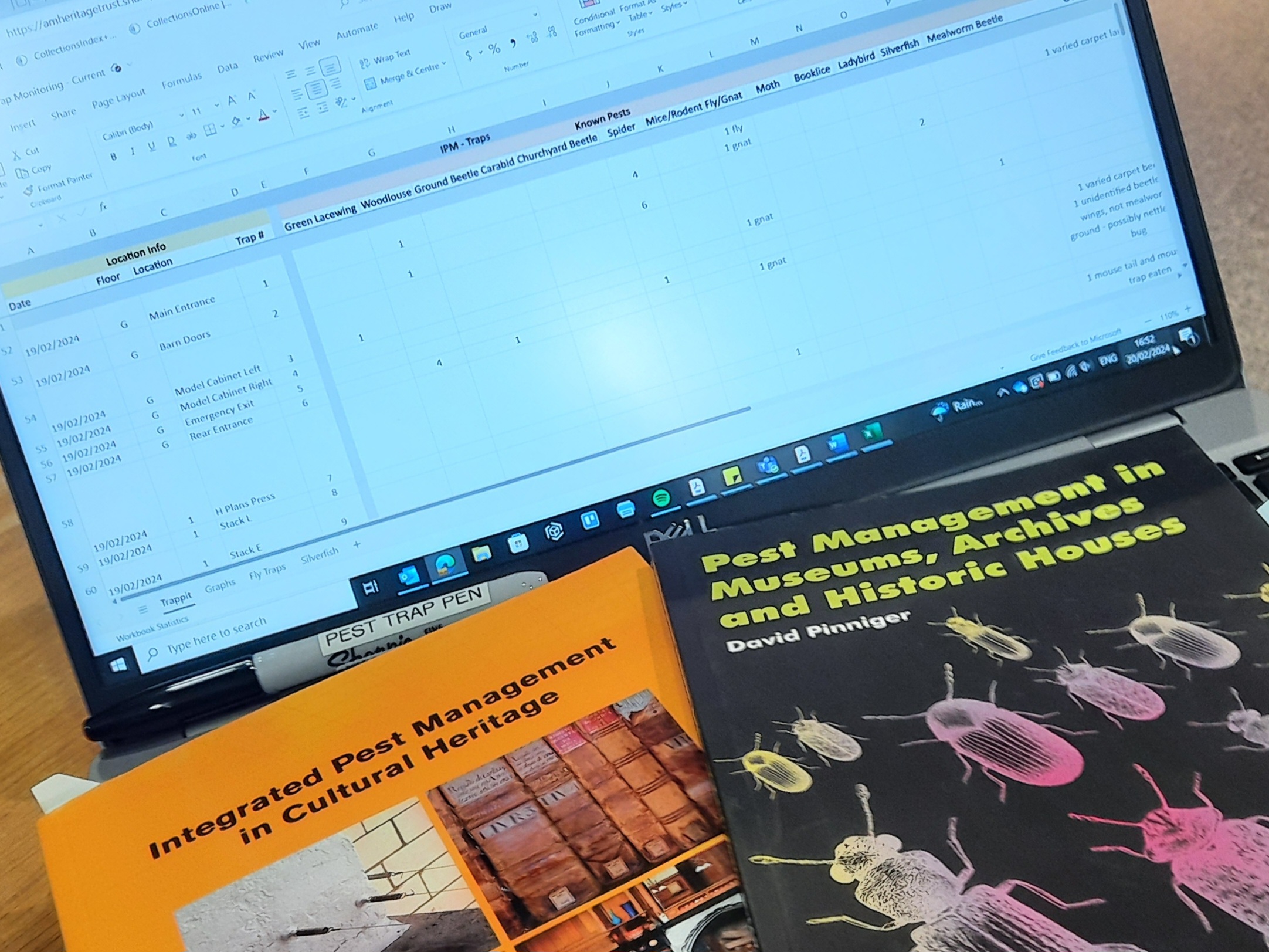Image shows a laptop open with a spreadsheet showing different types of insect pest species and the quantity of them found in different locations. Infront of the laptop are two books, Integrated Pest Manangement in Cultural Heritage and Pest Management in Museums, Archives and Historic Houses by David Pinniger. There is also a sharpie labelled 'pest trap pen'.