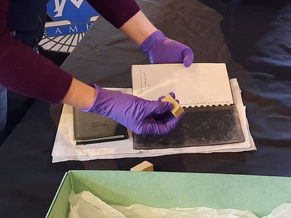 Cleaning a black mouldy book cover with visible white mould. The conservator holds a small piece of smoke sponge in her hand for the cleaning