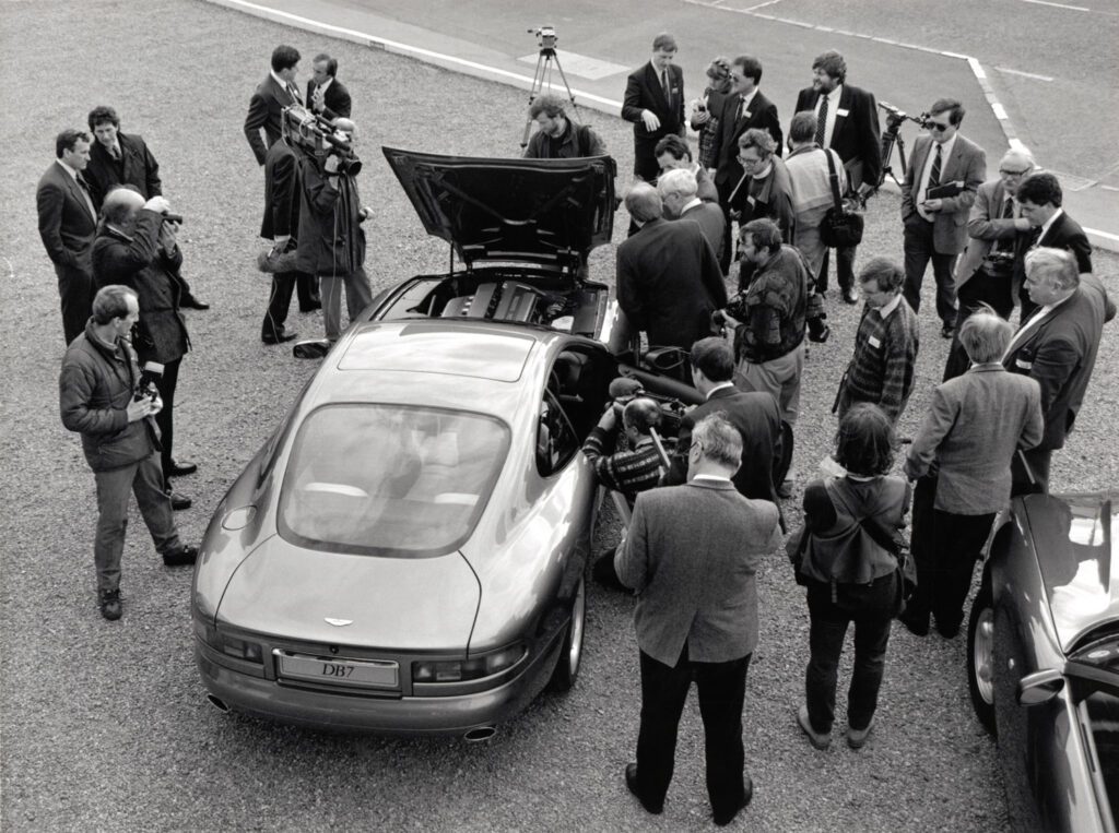 Black and white image of DB7 i6 prototype surrounded by press photographers and journalists