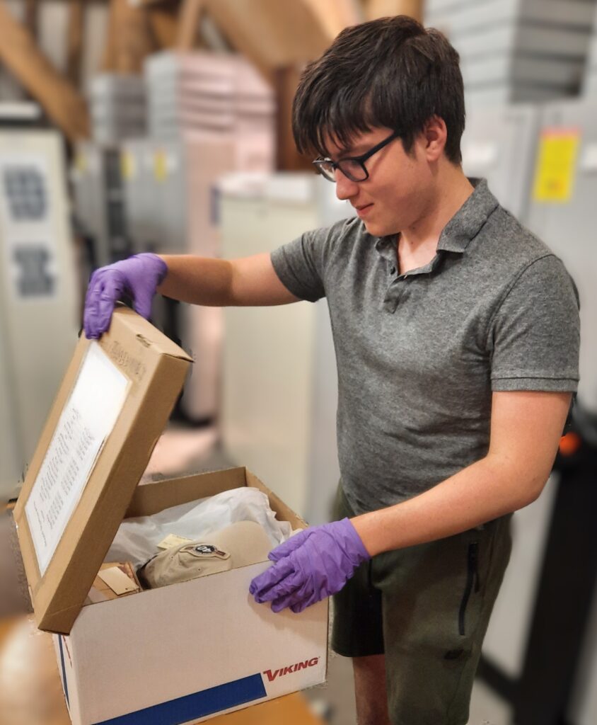 A young caucasian man with glasses and dark hair lifts the lid on a box of objects