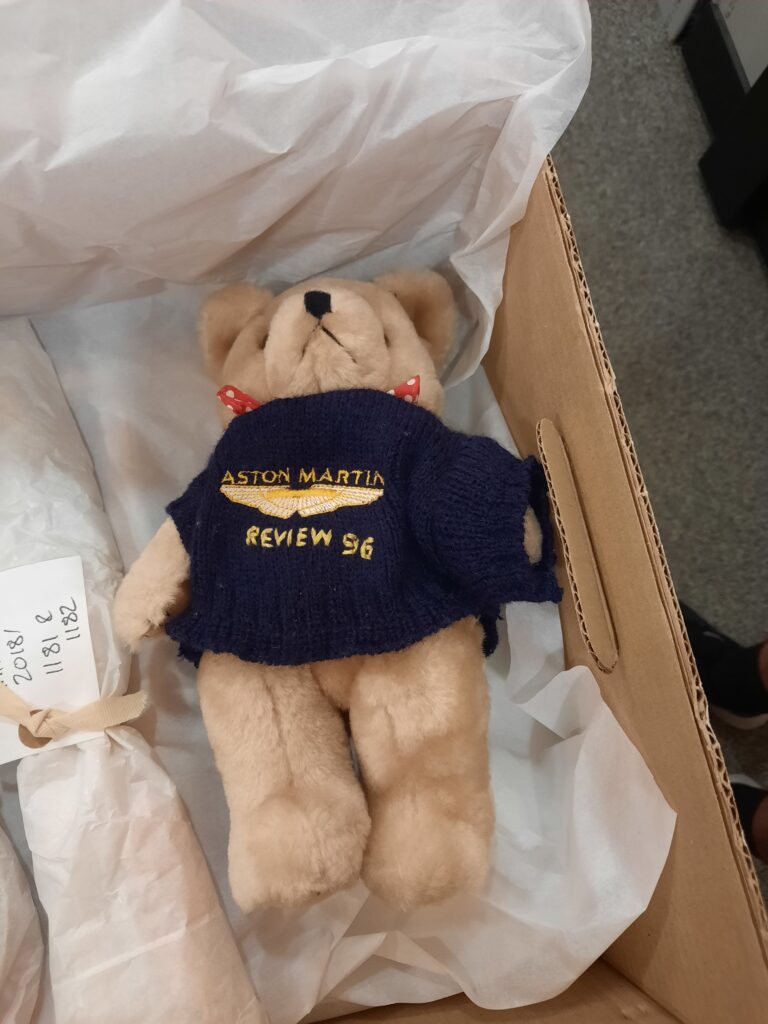 Teddy bear with a blue jumper on which is embroidered with the Aston Martin wings, Aston Martin and Review 96. The bear also has a red and white spotted neck tie sticking out above the jumper