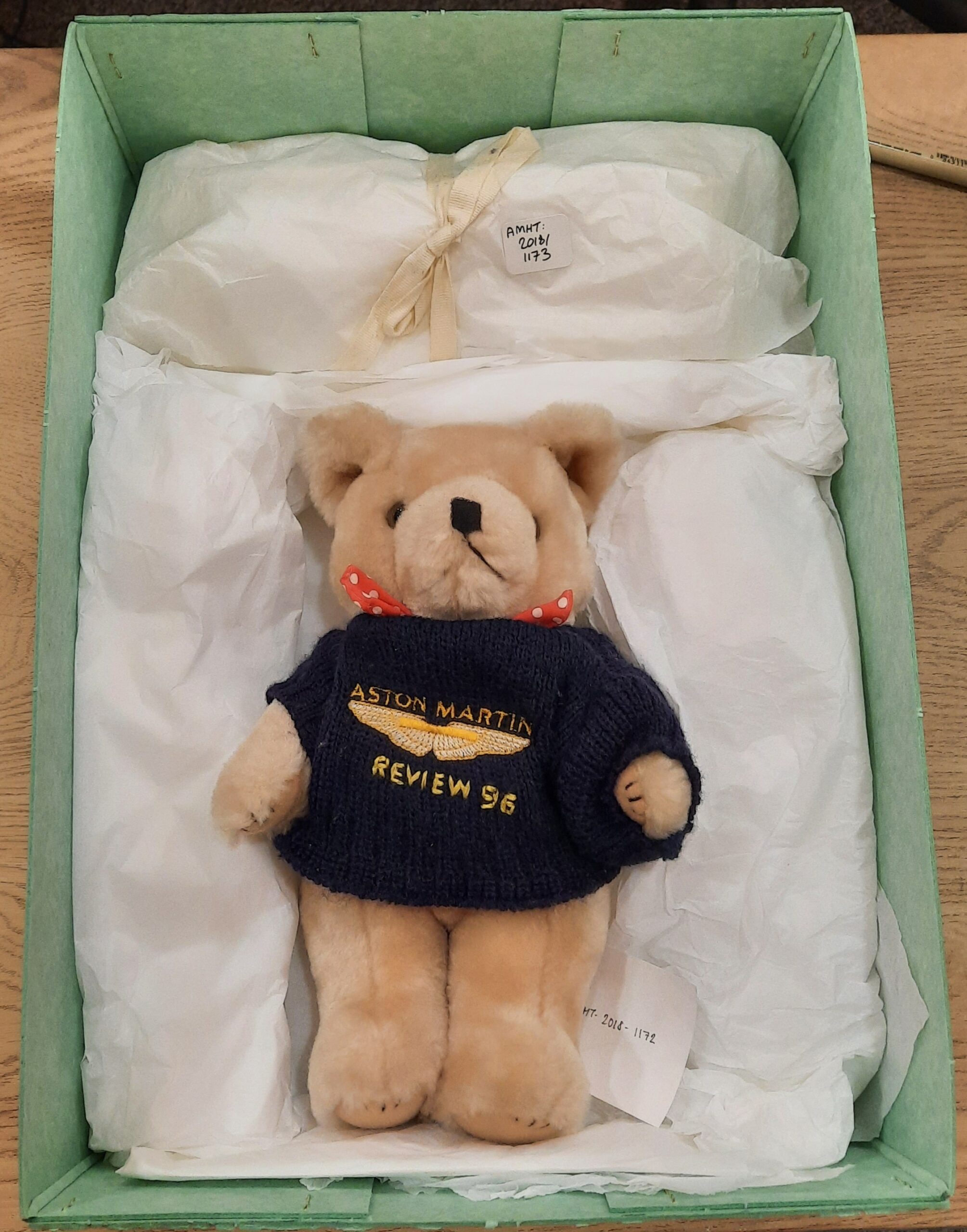 Teddy bear with a blue jumper on which is embroidered with the Aston Martin wings, Aston Martin and Review 96. The bear also has a red and white spotted neck tie sticking out above the jumper. The bear is laying in a green box, surrounded by tissue puffs