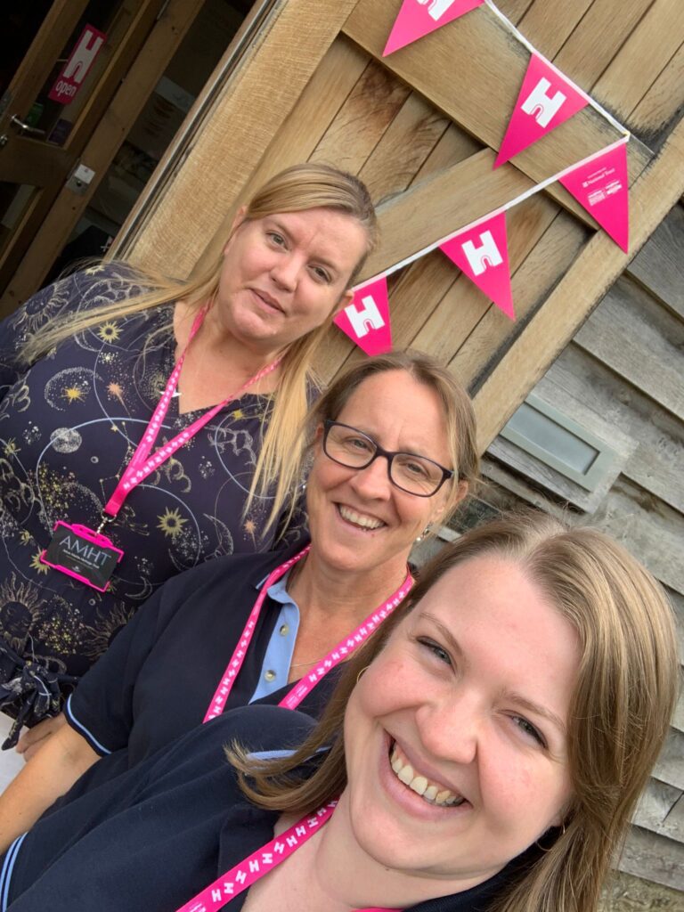 Image shows three Caucasian women with blond hair wearing pink Heritage Open Day lanyards in front of a wood panel door covered with pink bunting.