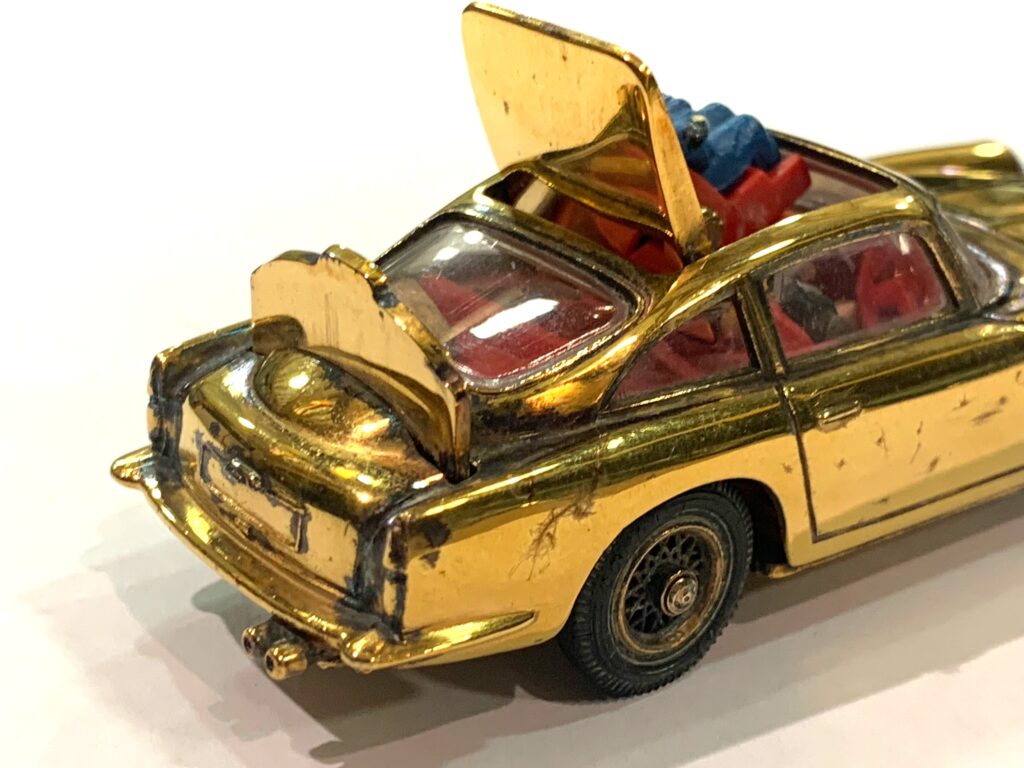 Image shows a close up of the rear of a gold coloured Corgi model of a DB5. It has the sun roof open so the 'baddy' is visible and the ejector seat up, it also has the rear bullet proof shield raised