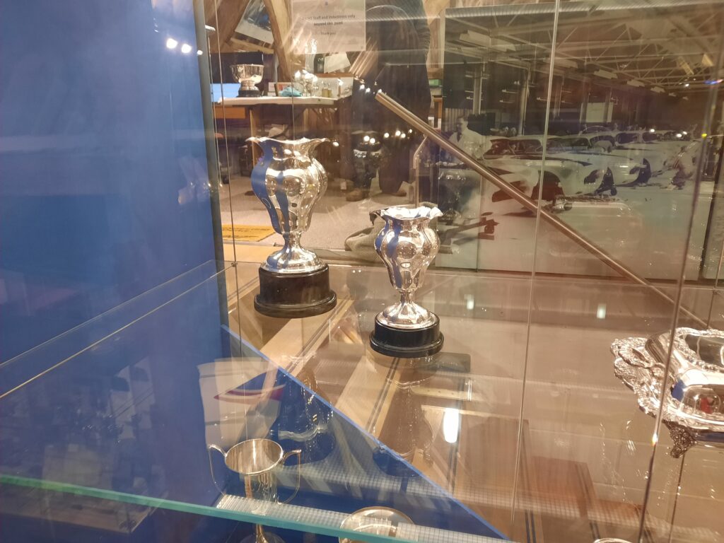 Two silver trophies on black plinths are displayed in a glass, see-through case with a dark blue wall to the left. In the background is a black and white canvas print which shows Aston Martin's being worked on in one of the early factories