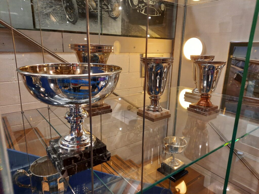 Four silver trophies on marble plinths are displayed in a glass, see-through case. Behind them aspects of artworks showing Aston Martins can be seen.