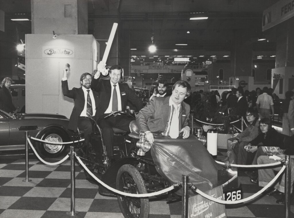 Black and white image of three AML employees, including company directors, smiling at the camera sitting in an old Lagonda bicycle style 'vehicle'.