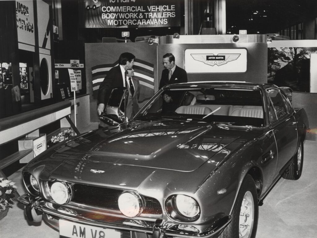 Black and white image of Aston Martin's display stand at a motor show at the NEC in Birmingham. A V8 Vantage is on display with three people stood behind the car with the drivers side door open