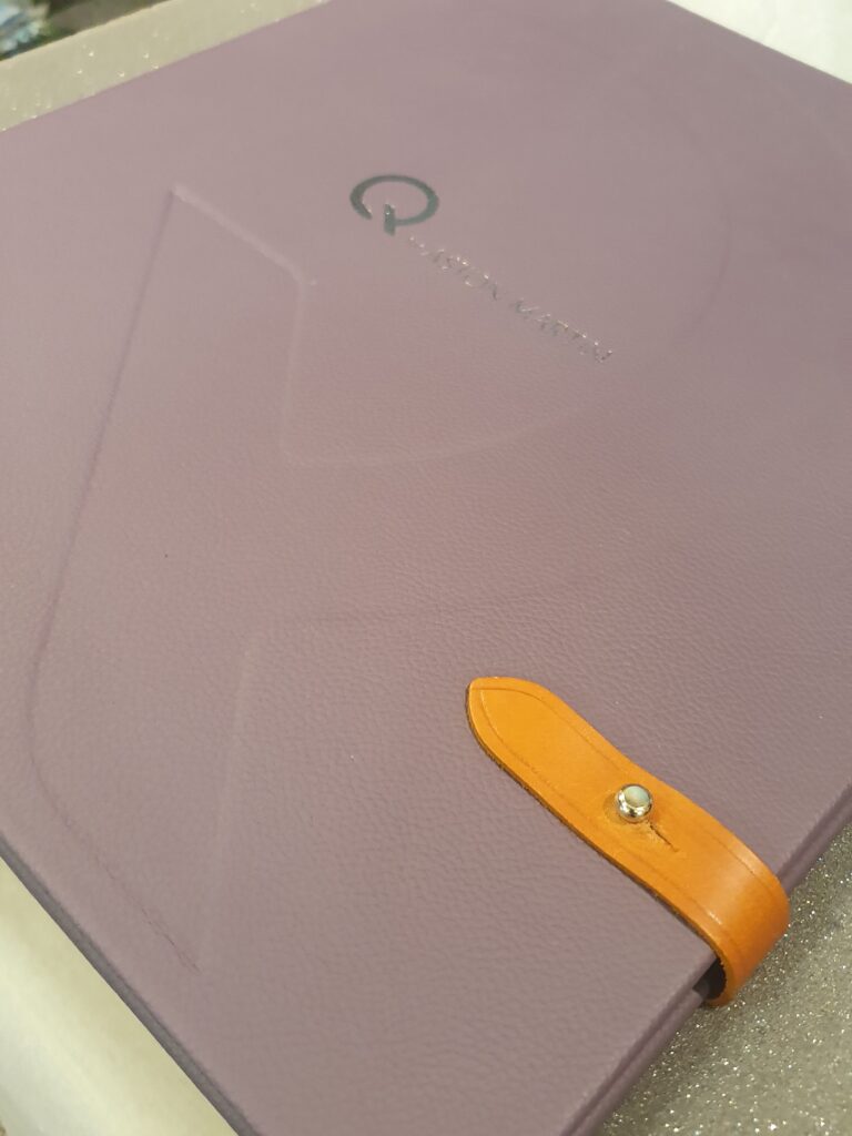 Image shows an angled close-up of a purple leather bound large book with tan leather straps folded over the right lower side to seal it. On the front is Q by Aston Martin in silver. A large Q imprint is stamped on the brochure.