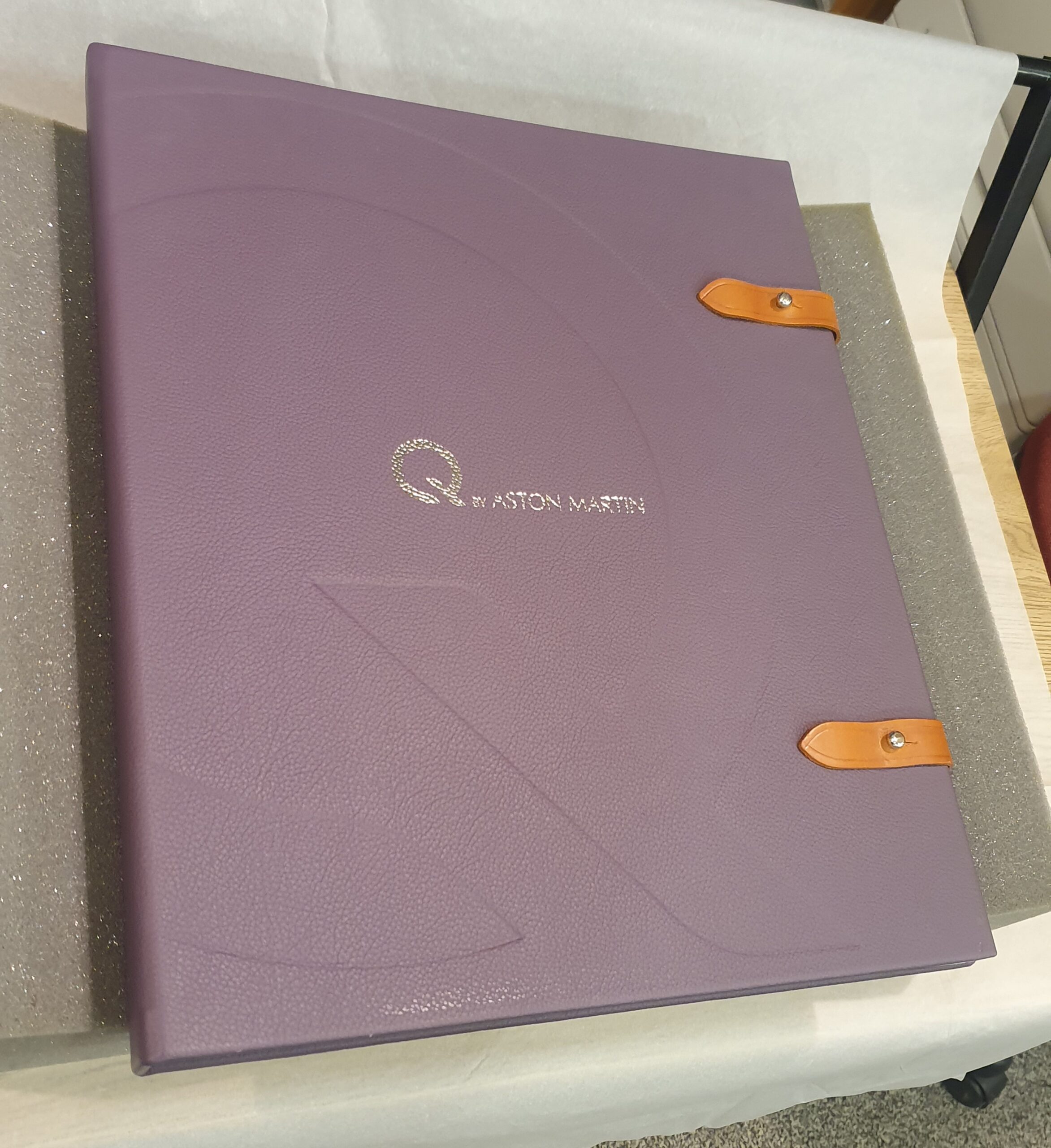 Image shows purple leather bound large book with tan leather straps folded over the right edge to seal it. On the front is Q by Aston Martin in silver. A large Q imprint is stamped on the brochure.