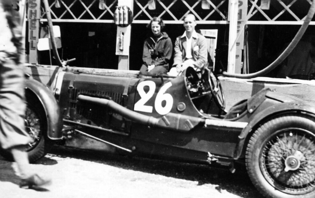 Black and white image of Aston Martin LM7, with race number 26 on the side, and two racing drivers sitting on the pit wall behind it. Elsie 'Bill' Wisdom on the left, a Caucasian woman in dark overalls, and Mort Morris-Goodall, a Caucasian man in white overalls on the right.