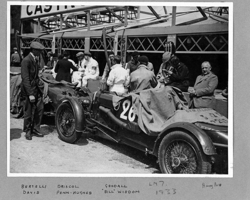 Black and white image of Aston Martin LM7, race number 26, in the pit lanes surrounded by drivers in overalls and flat caps