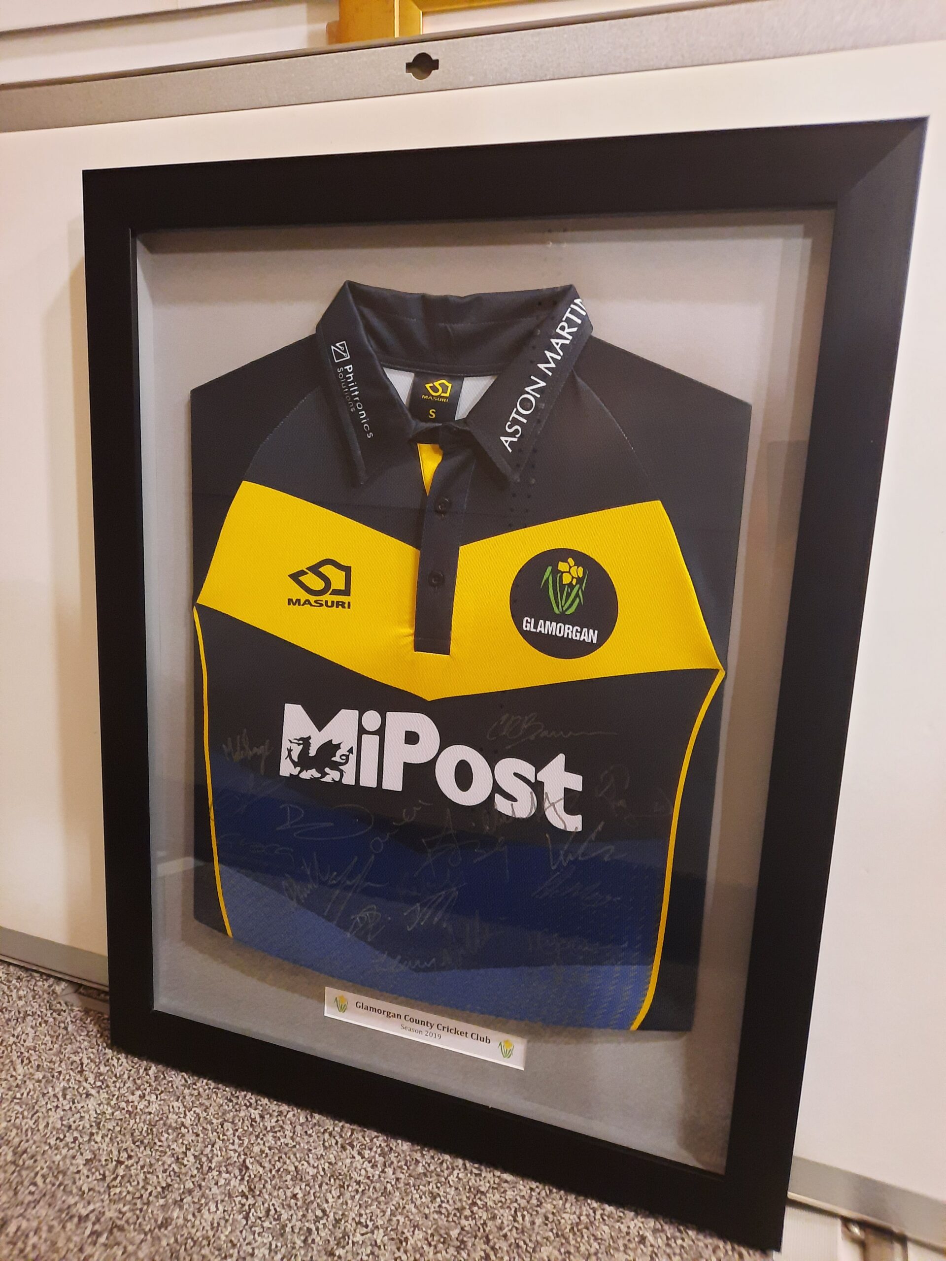 Framed dark blue and yellow cricket shirt for the Glamorgan County Cricket Club first team. The shirt has a yellow stripe across the top of the chest, with a daffodil within a black circle on the left breast. Sponsors logos are in white across the chest on the collar.
